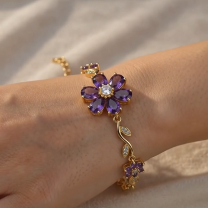 A gold-tone bracelet with a purple flower centerpiece and small clear stones, worn on a person's wrist against a soft beige fabric background.